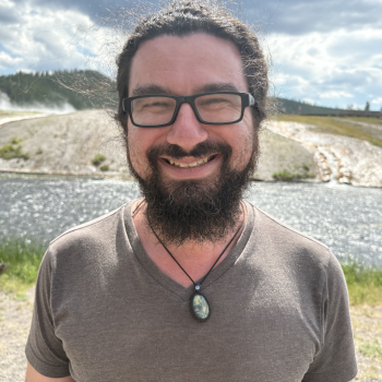 A smiling man with a beard and glasses stands outdoors near water wearing a brown shirt and pendant necklace.