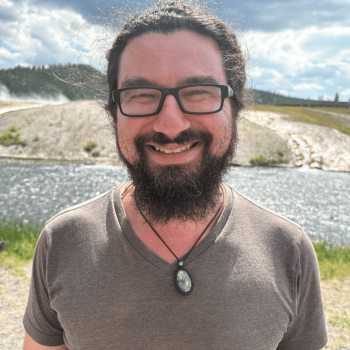 A smiling man with a beard and glasses stands outdoors near water wearing a brown shirt and pendant necklace.